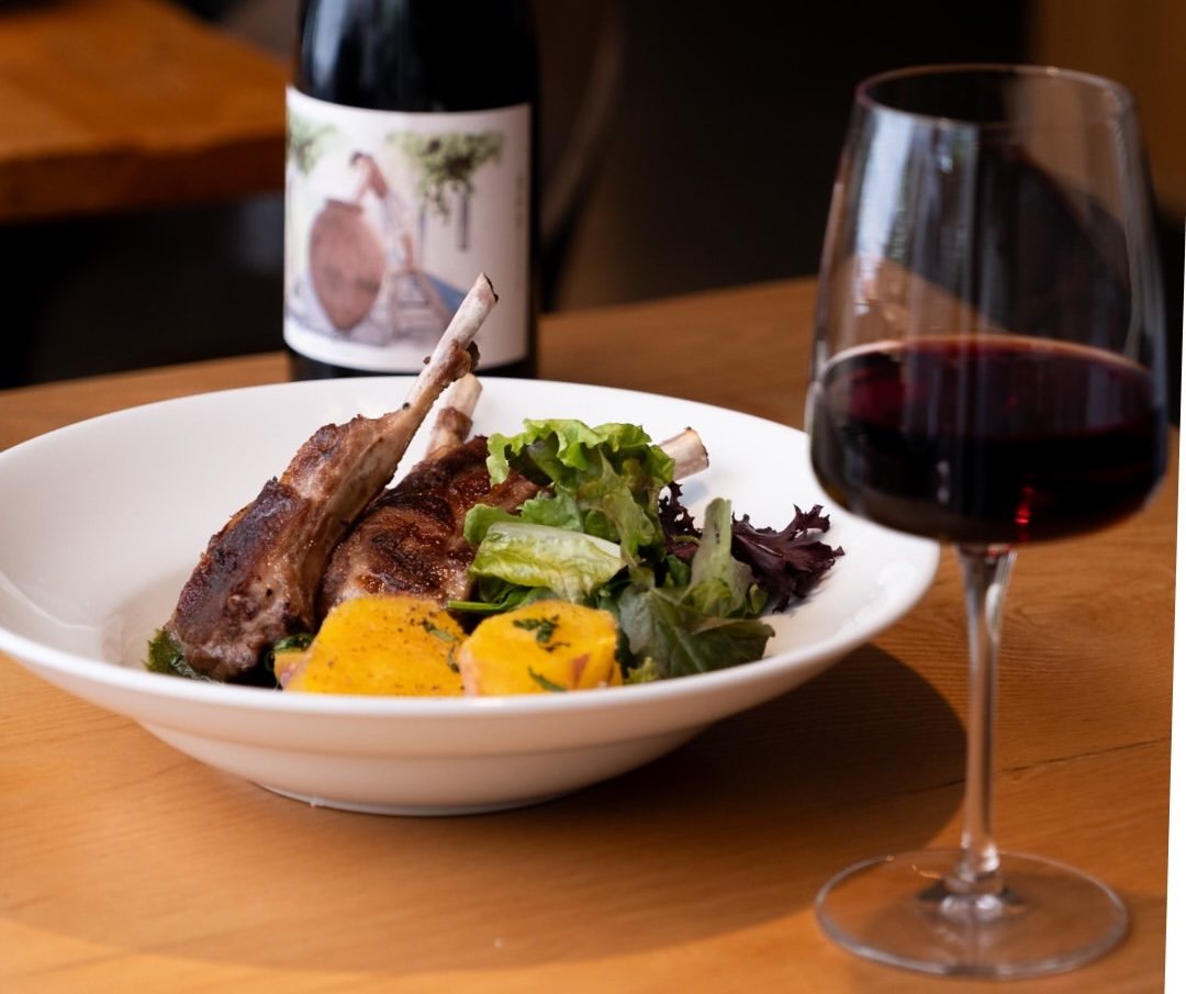 Prato fundo com borrego e salada, ao lado de um copo de vinho tinto